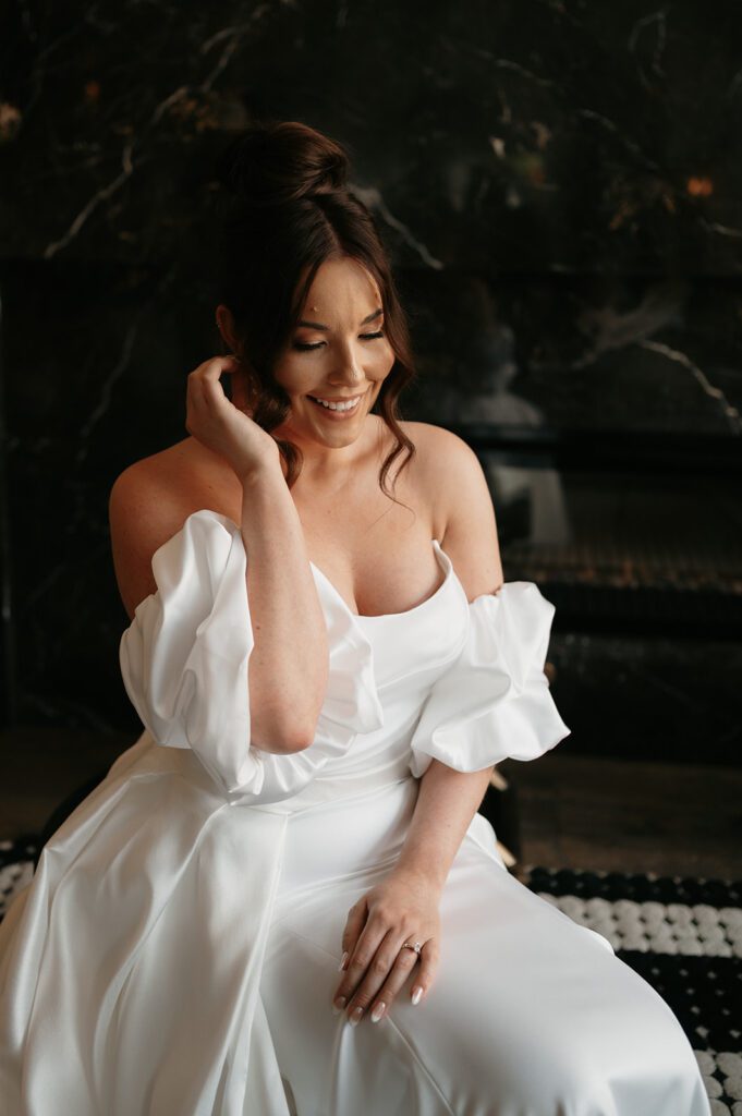 A Des Moines bride on her Iowa wedding day, styled by Hello Darlin' Glam, smiles radiantly while sitting in her white off-the-shoulder wedding gown with ruffled sleeves. Her elegant updo with soft, loose curls framing her face complements her glowing makeup, perfectly highlighting her natural beauty. She gently rests one hand near her ear, exuding a calm and joyful expression. The dark marble background adds contrast, making her bright bridal look stand out.